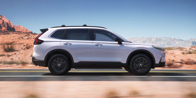 A white SUV with black roof racks drives right on a paved road through a desert landscape with mountains.
