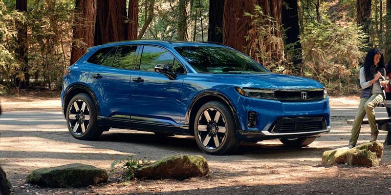 Blue Honda Prologue electric SUV parked in a redwood forest setting.